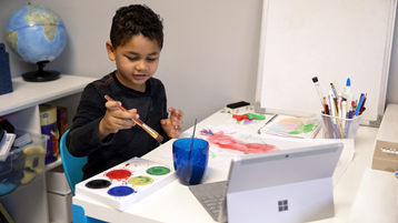 Ένα νεαρό αγόρι χρησιμοποιεί χρώματα σε χαρτί καθώς παρακολουθεί έναν ανοιχτό φορητό υπολογιστή Surface