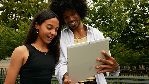 A young man shows a young woman something on a Surface Pro device in a park setting.