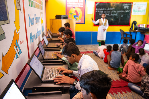 An educator presenting in a school in India while students work on laptops.