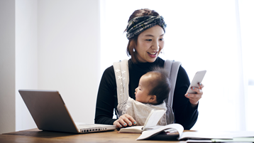 A smiling Japanese woman holds her baby in a carrier as she checks her phone and works from a laptop