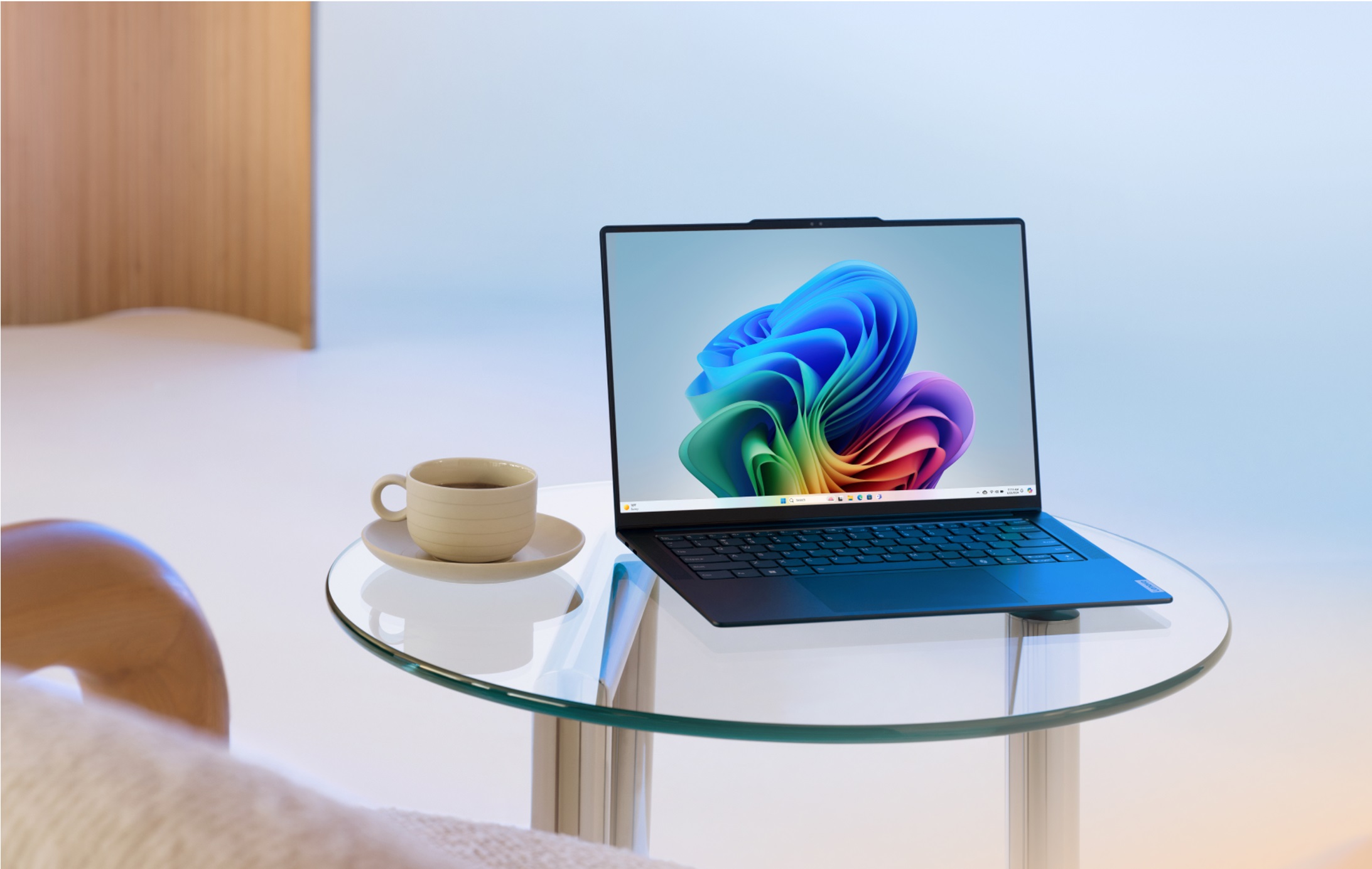 Un PC Windows 11 mostrando el fondo de escritorio de floración multicolor. El portátil está sobre una pequeña mesa circular de cristal junto a una taza de café.