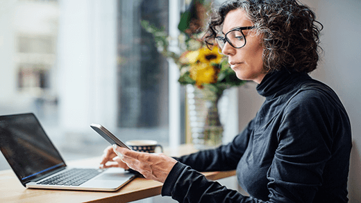 Empresaria madura sentada en un café mirando su teléfono móvil mientras trabaja en un equipo portátil. Mujer leyendo un mensaje de texto en una cafetería.