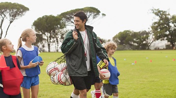 kuva lasten urheiluvalmentajasta kantamassa varusteita pelikentälle