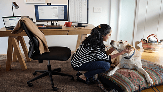 אישה עובדת מרחוק מהמשרד שלה בבית, לוקחת הפסקה ומלטפת את הכלב שלה; מכשיר Latitude 13 של Dell.
