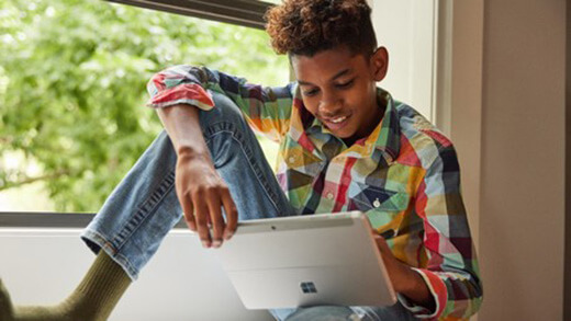 Seorang siswa laki-laki muda duduk di jendela melihat perangkat Surface Pro.