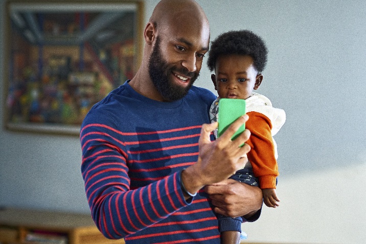 Fotoattēls ar vīrieti, kas tur mazu bērnu un sarunājas tālrunī
