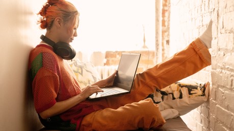 Młoda kobieta z pomarańczowymi włosami siedzi wygodnie w pobliżu okna ze słuchawkami nausznikami na szyi, patrząc na jej Windows 11 laptopa.