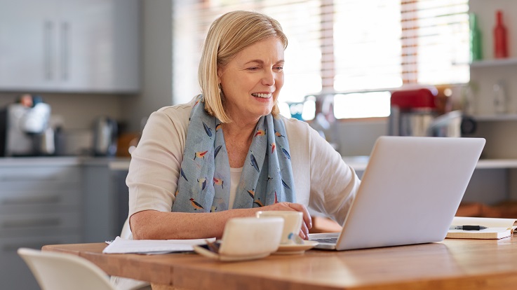 fotografie cu o femeie la o masă de bucătărie, uitându-se la e-mailuri pe un computer