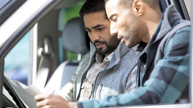 Dva čoveka gledaju u papirne zadatke – jedan čovek sedi u vozaču automobila automobila sa automobilom, a drugi pored njega