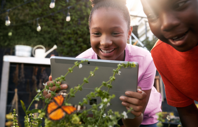 在花园里的K-12女学生拿着 Surface Go，同时拍摄蝴蝶的照片。 微笑的男K-12学生站在她旁边。
